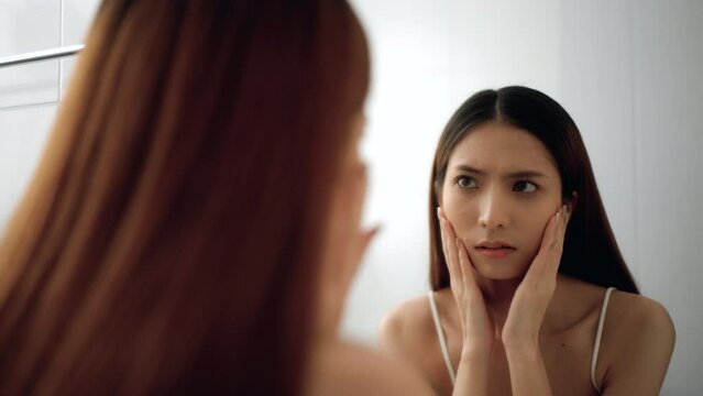 Worry feeling of asian woman touching unhealthy face skin looking at mirror and checking dry irritated face skin in bathroom. Unhappy Young female looking at red acne spots on face skin care.