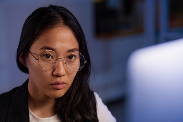 Close-up portrait of Asian female programmer wearing eyeglasses working on code at workplace in IT company office late in evening