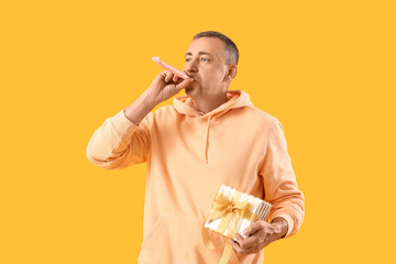 Mature man with party blower and birthday gift on orange background