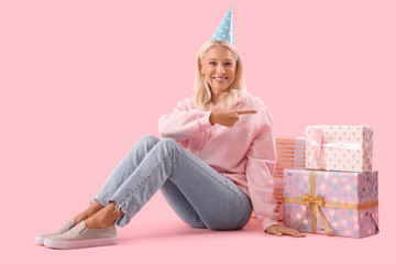 Happy mature woman in party hat pointing at gift boxes on pink background