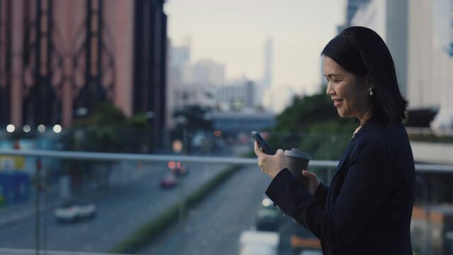 Side View of the Smiling Asian Woman Walking on the Bridge, Holding the Cup of Coffee Using the Cell Phone. Busy Happy Business Person Going to Workplace and Scrolling the Social Media Outdoors.