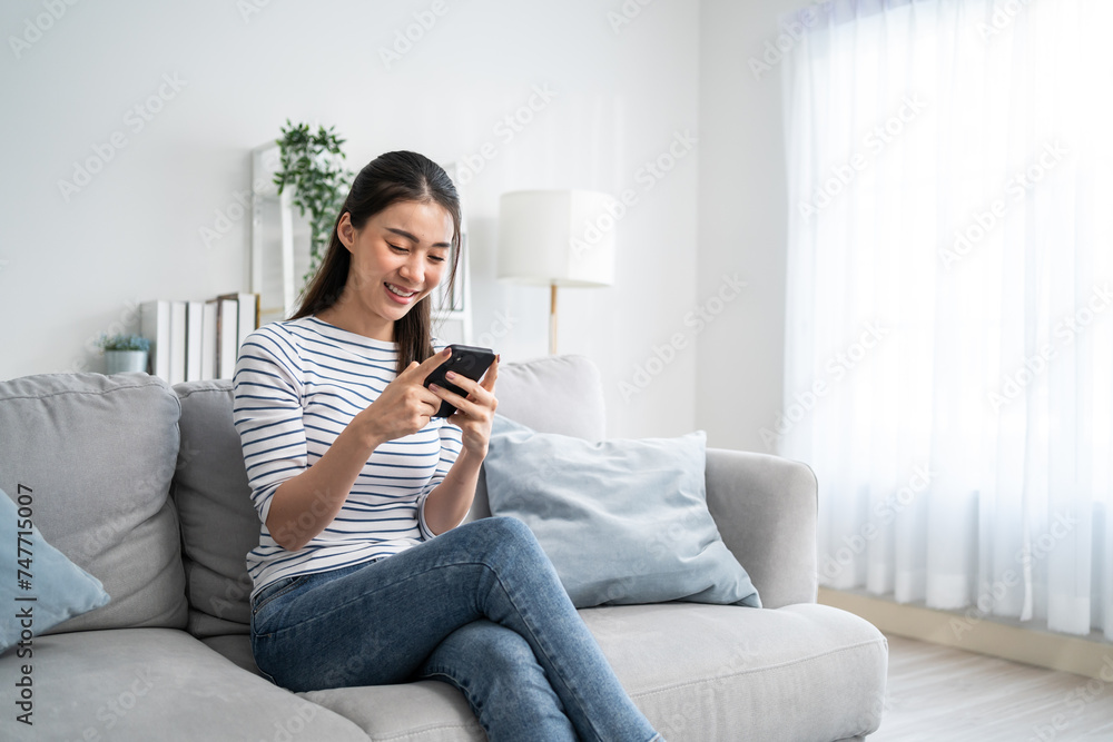 Wall mural asian beautiful woman sitting on sofa and swipe mobile phone in house.