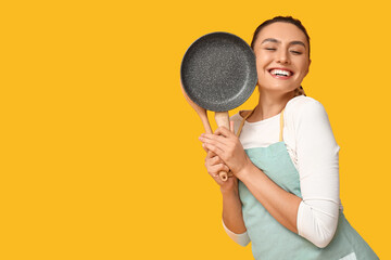 Portrait of happy young housewife in apron with frying pan and wooden spatula on yellow background