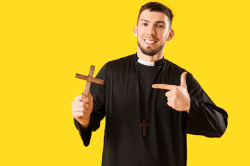 Young priest pointing at cross on yellow background