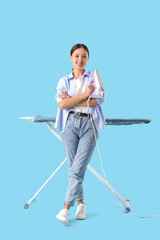 Happy Asian woman with iron standing near ironing board on blue background