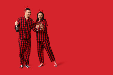 Happy young couple in Christmas pajamas and with champagne on red background