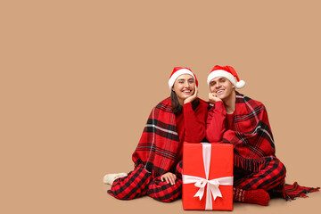 Happy young couple in Santa hats, with plaid and Christmas gift on beige background