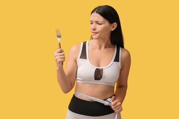 Beautiful young displeased woman with fork and measuring tape on yellow background. Weight loss concept