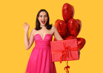 Beautiful young happy woman with gift box and heart-shaped balloons on yellow background....