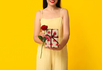 Beautiful young woman with gift box and red rose on yellow background. Valentine's Day celebration