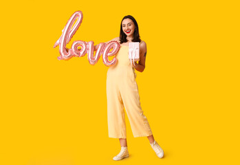 Beautiful young happy woman with gift box and balloon in shape of word LOVE on yellow background....