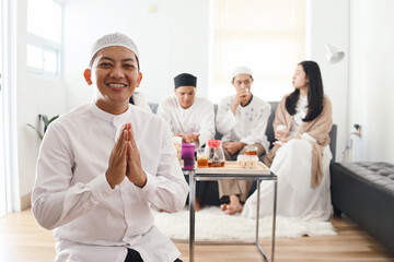 Friendly Asian muslim man wear skullcap showing greeting or welcome gesture during Eid mubarak celebration and his friends gather on the background - obrazy, fototapety, plakaty