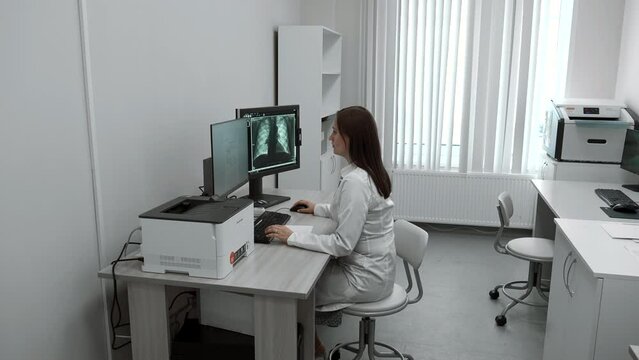 Female doctor looks at X-ray on computer. Clip. Doctor in bathrobe looks at tests and X-rays in computer room. Modern electronic technologies in medical clinics