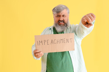 Protesting mature man holding placard with word IMPEACHMENT and pointing at viewer on yellow...