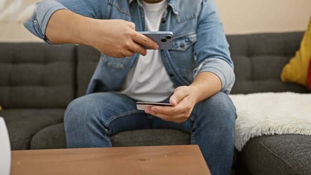 Man using smartphone and holding credit card for online payment while sitting on couch indoors.