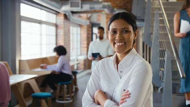 Portrait of smiling young businesswoman working in modern open plan office turning to look at camera and folding arms with colleagues in background - shot in slow motion