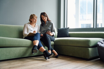 Two business women sitting on sofa smiling while using digital tablet during meeting in office