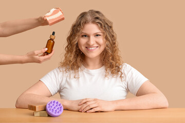 Beautiful young woman with hair scalp massager and hands holding cosmetic products on beige background