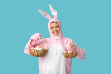 Beautiful young woman in bunny costume holding wicker baskets with Easter cake and eggs on blue background