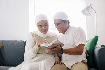 Asian Muslim man teaching woman reading Koran or Quran in living room. Muslim couple praying on the couch at home. - obrazy, fototapety, plakaty