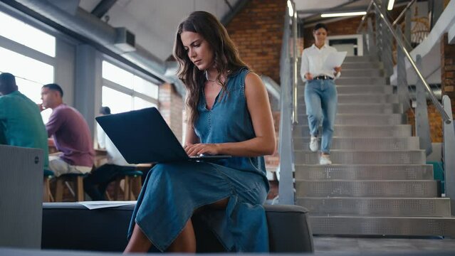Young businesswoman with serious expression working on laptop computer in modern open plan office with colleagues in background  - shot in slow motion