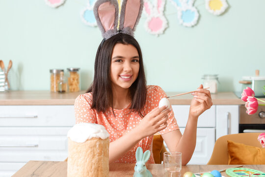 Beautiful young woman in bunny ears painting Easter egg in kitchen