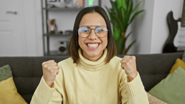At home, a young and beautiful hispanic woman ecstatically screaming in victory, her arms raised high. wearing glasses, she celebrates her success with unadulterated joy and pride, exuding positivity.