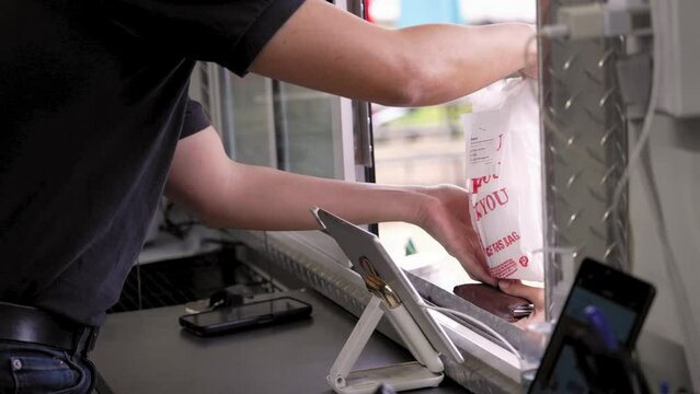 Worker hands prepared packaged food order through food trailer truck window to customer holding wallet, slow motion 4K