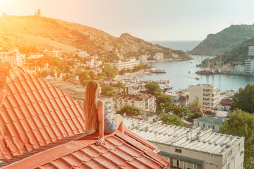 Woman sits on rooftop, enjoys town view and sea mountains. Peaceful rooftop relaxation. Below her,...