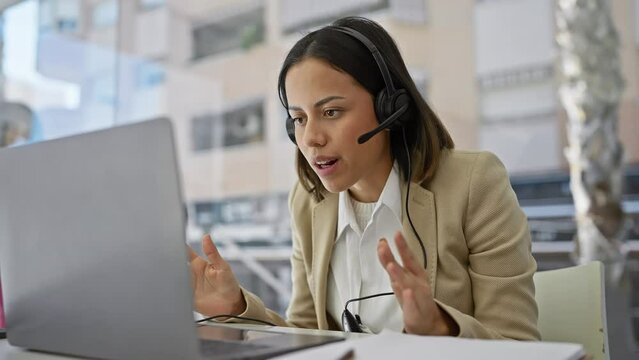 Young hispanic woman with headset working on laptop in modern office with city view.