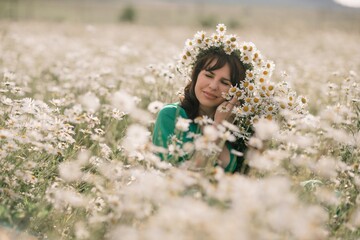 Happy woman in a field of daisies with a wreath of wildflowers on her head. woman in a green dress in a field of white flowers. Charming woman with a bouquet of daisies, tender summer photo