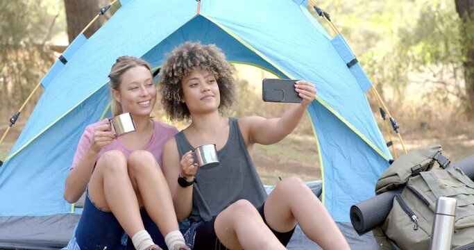 Two women sit inside a tent, one holding a mug and the other taking a selfie