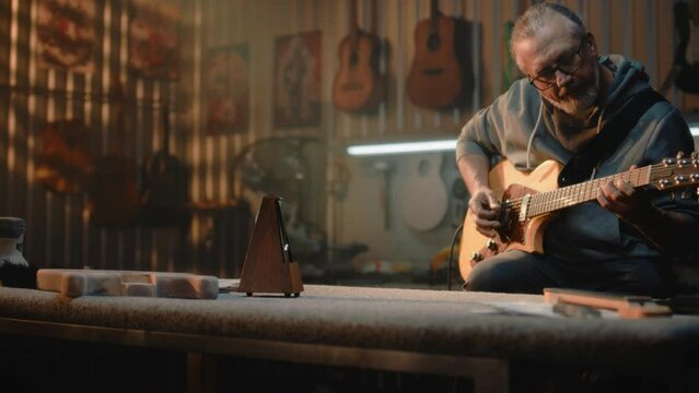 Mature musician or professional artisan plays guitar in stylish workshop for making musical instruments. Camera changes view to metronome standing on table. Handmade wooden guitars hang on the wall.