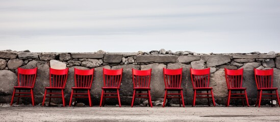 A row of red chairs lined up neatly next to a sturdy stone wall, creating a simple and inviting outdoor seating area. - Powered by Adobe