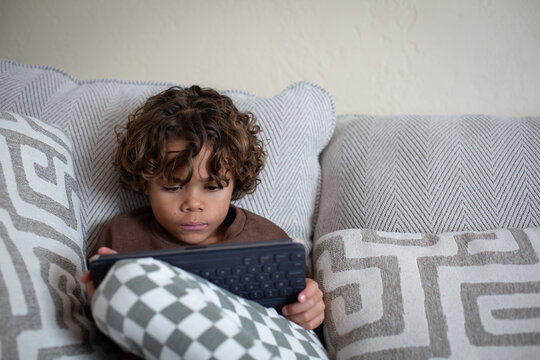 Young diverse little boy playing on a tablet computer sitting on the couch in his home. Spending his free time staring at a screen and playing video games