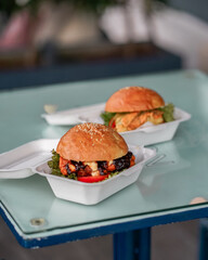 A close-up of two hamburgers with cheese, vegan, meat, and eggs in styrofoam containers on a table