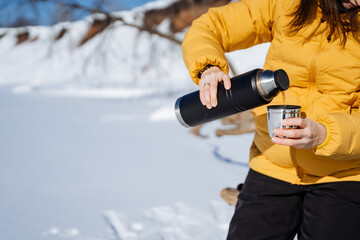 Hiking in the mountains, woman holding a thermos with tea in her hands and pouring a drink into a mug against the background of snow, winter trekking alone, warming drink.