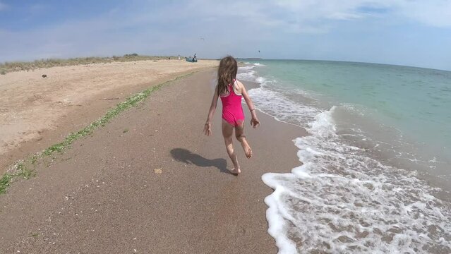 Happy playful young girl, kid running barefoot in pink swimwear on sandy beach in summer vacation, sea waves approaching the shore