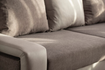 The sofa is a combination of light mottled and gray with shadows and sun rays. Interior upholstered furniture with a wide armrest and pillows for relaxation with a blurred background close-up.