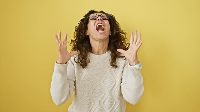 Furious middle age hispanic woman in glasses, going berserk, standing, yelling in deep frustration, arms raised madly. isolated yellow backdrop screaming deed.