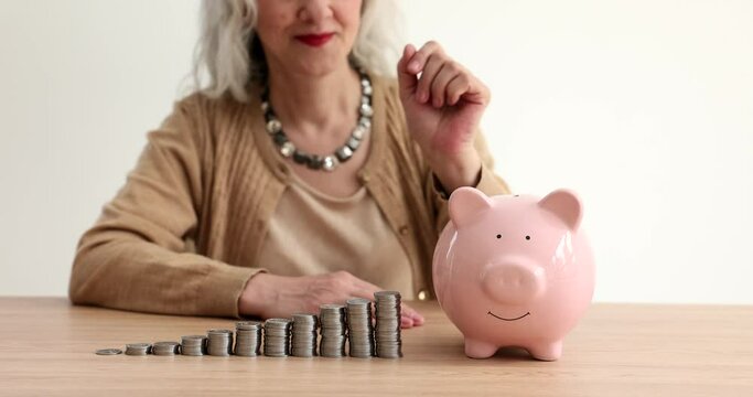 Elderly woman throwing coins into piggy bank near piles of kopecks in ascending order closeup 4k movie slow motion. Pension increase reform concept
