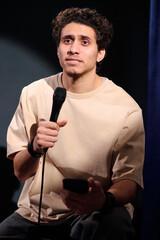 Young comedian of stand up club speaking in microphone and using smartphone during monologue in...