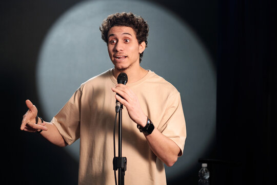Young comedian of stand up club standing on stage in front of audience and pronouncing monologue in microphone during performance
