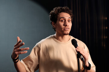 Young male comedian of stand up club gesticulating and speaking in microphone while pronouncing...