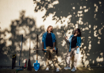 Two happy lesbian girls pose and laugh with garden tools near the wall in the shade of trees and...