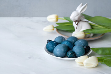 Aesthetic spring traditional Easter holiday table decoration, plate with blue colored eggs, bunny figurine, white tulip flowers on neutral marble tabletop background, soft selective focus