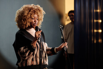 Young African American female presenter or comedian in wavy wig speaking in microphone during...