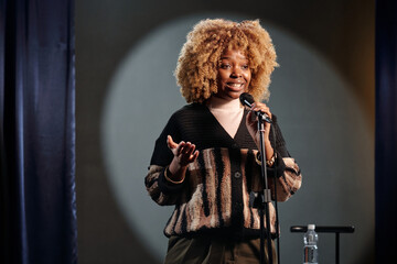 Young African American stand up comedian in wig speaking in microphone during performance while...