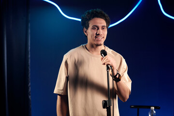 Young male comedian in beige t-shirt speaking in microphone on stage while looking at audience while pronouncing monologue - obrazy, fototapety, plakaty
