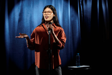 Young comedian in eyeglasses and brown shirt standing on stage against blue curtains and speaking in microphone during performance